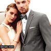 A handsome man wearing the grey 'Affection' Tuxedo by Michael Kors stands in front of a helicopter with a beautiful woman in his arms.