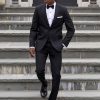 A handsome black man wearing the black 'Quincy' tuxedo walks down a flight of concrete stairs in front of a gorgeous mansion with his hand causally in one pocket.